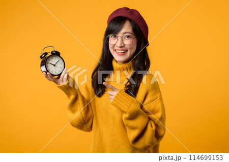 In a cheerful scene, a young woman in a yellow sweater and red beret shows off her alarm clock, embodying punctuality and success. 114699153