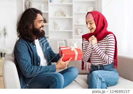 Eastern man and a woman are sitting on a couch, both holding a present. They seem to be in a cozy living room setting, sharing a moment of gift-giving and celebration. 114699157