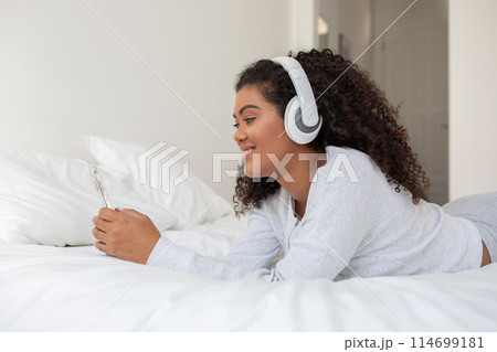 A young Hispanic woman with curly hair lies comfortably on a white bed, dressed in a casual white outfit, enjoying music through her white headphones, with a smartphone in her hand. A young Hispanic woman with curly hair lies comfortably on a white bed, dressed in a casual white outfit, enjoying music through her white headphones, with a smartphone in her hand. 114699181