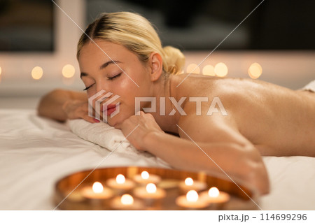 A woman is laying on top of a bed next to a row of burning candles at spa. The room is dimly lit, casting a warm glow on the scene. 114699296