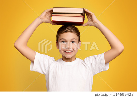 Positive schoolboy holding stack of books on his head, orange studio background 114699428