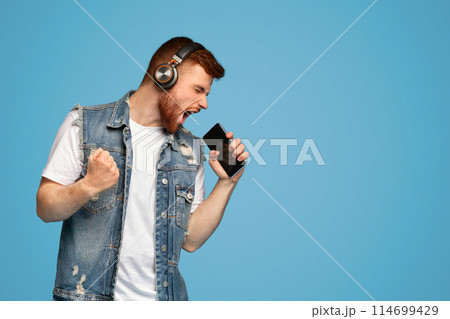Young emotional millennial redhaired guy singing his favorite song on smartphone as microphone on blue background, copy space 114699429