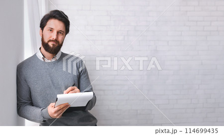 Waiting for patient. Friendly psychologist standing with clipboard, leaning at wall in office, empty space 114699435