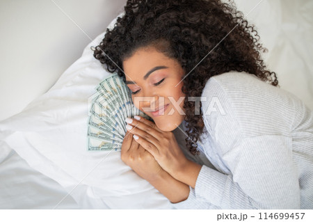 A Hispanic woman is seen laying on a bed with a large amount of money spread out around her. She appears to be counting or looking at the money. 114699457