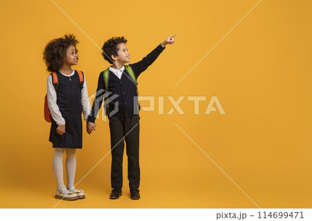African American young boy and a girl schoolers are pointing at an indiscernible object or direction on yellow background. Their fingers are extended and they are looking intently at the same point. 114699471