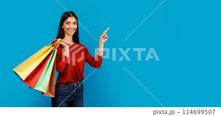 A cheerful young woman dressed in a casual red top and blue jeans stands against a solid blue backdrop, holding colorful shopping bags in one hand while pointing at copy space A cheerful young woman dressed in a casual red top and blue jeans stands against a solid blue backdrop, holding colorful shopping bags in one hand while pointing at copy space 114699507