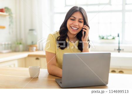 A middle eastern woman is multitasking, talking on her cell phone while working on her laptop. She appears focused and engaged in a conversation while typing on the computer. 114699513