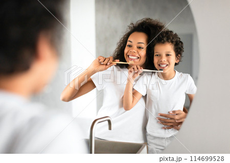 African American woman is standing next to a child, brushing her teeth with a toothbrush in a bathroom. The child is looking up at the woman, potentially learning how to brush his own teeth. 114699528