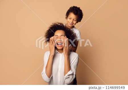 African American young child is gleefully covering his mothers eyes from behind as they both share a moment of joy and surprise 114699549