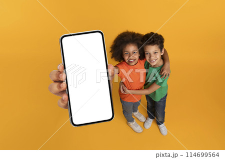 African American kids holding up a cell phone with a blank white screen, showing no content or images, mockup copy space, yellow studio background 114699564