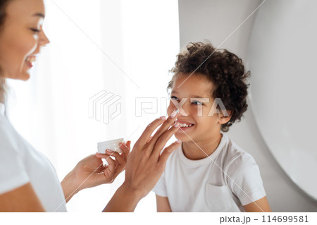 A joyful African American boy with curly hair sits indoors as a smiling woman tenderly applies a dab of cream to his nose. The casual, affectionate moment captures a daily skincare routine 114699581