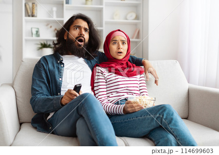 Indian man and woman are sitting side by side on a white couch in a well-lit living room. Both appear surprised and shocked by something they see on the television screen. 114699603