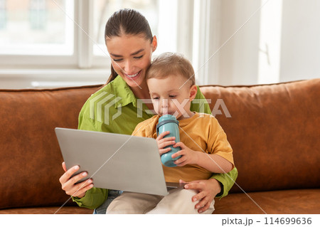 A woman sits on a couch with a child next to her, both engaged with a laptop. Mother appears to be typing, while her son looks on attentively. 114699636