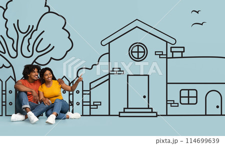Happy black couple are seated on the ground, facing a detailed drawing of a house. The drawing appears to be on a large sheet of paper laid out on the ground 114699639