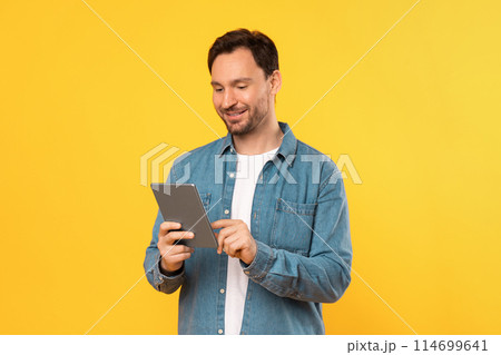 Guy wearing a blue shirt is engrossed in using a tablet device. He is focused on the screen, possibly reading or viewing content 114699641