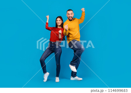 A man and woman exude happiness and success as they pump their fists in the air against a bright blue backdrop 114699683