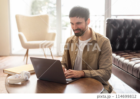 A man sits at a table, focused on his laptop computer. He appears to be working or studying intently, with papers and a drink nearby. 114699812