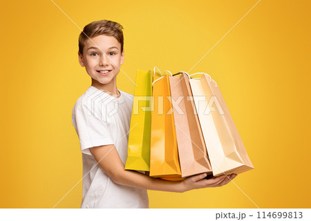 Little shopper. Happy teen boy holding lots of colorful paper shopping bags in hands, orange studio background 114699813