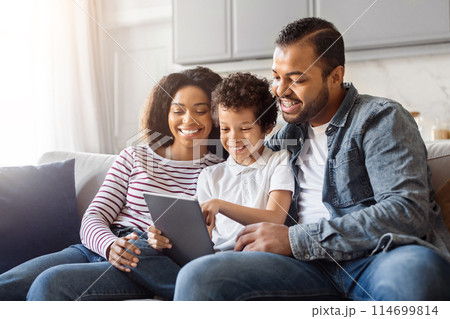 African American man, woman, and child are sitting together on a couch, engrossed in looking at a tablet. The man holds the device, while the woman and child lean in, focusing on the screen. 114699814
