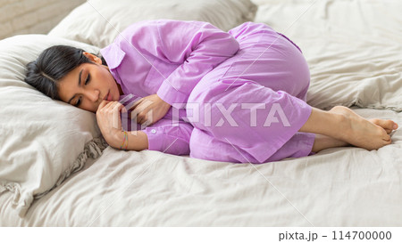 Depressed young middle eastern woman is curled up in a comfortable position, wearing soft purple pajamas, on a plush white bed Depressed young middle eastern woman is curled up in a comfortable position, wearing soft purple pajamas, on a plush white bed 114700000