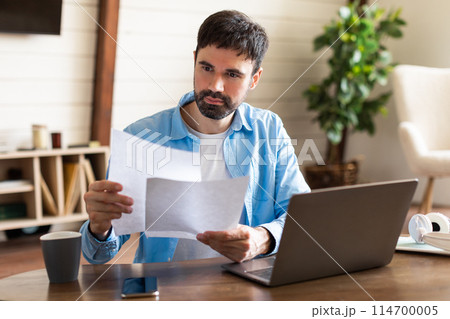 A man is standing and holding a piece of paper in front of a laptop. The man appears focused while looking at the laptop screen and holding the paper A man is standing and holding a piece of paper in front of a laptop. The man appears focused while looking at the laptop screen and holding the paper 114700005