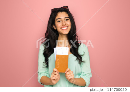 A cheerful young middle eastern woman stands against a vibrant pink backdrop, clutching a passport and a boarding pass, suggesting she is prepared and excited for an upcoming journey A cheerful young middle eastern woman stands against a vibrant pink backdrop, clutching a passport and a boarding pass, suggesting she is prepared and excited for an upcoming journey 114700020
