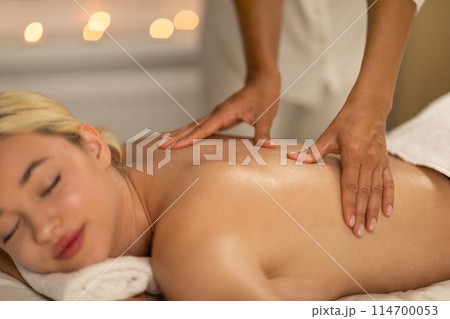 A woman is lying face down on a massage table while a massage therapist is giving her a back massage at a luxurious spa. The therapists hands are applying pressure to the womans muscles, closeup A woman is lying face down on a massage table while a massage therapist is giving her a back massage at a luxurious spa. The therapists hands are applying pressure to the womans muscles, closeup 114700053