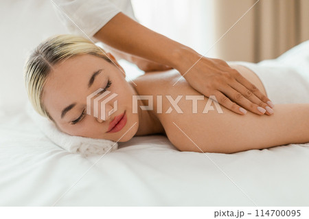 A woman lies face down on a massage table in a serene spa setting while a massage therapist performs a back massage on her. The therapists hands are visible as they apply pressure and knead A woman lies face down on a massage table in a serene spa setting while a massage therapist performs a back massage on her. The therapists hands are visible as they apply pressure and knead 114700095