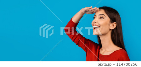 A joyful young woman wearing a bright red top is standing against a solid blue backdrop. She has her hand above her eyes as if shading them from the sun, gazing into the distance, copy space 114700256
