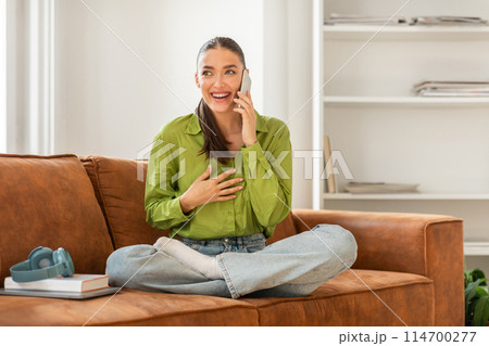 A woman is seated on a couch, engaged in a conversation on her cell phone. She appears focused on the call, with a concentrated expression on her face. 114700277