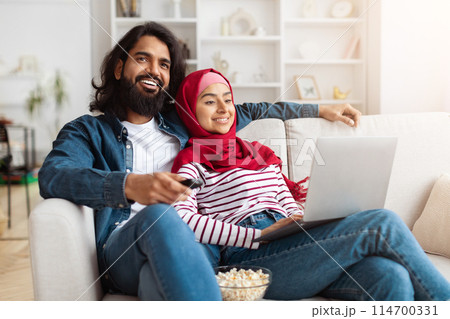 Muslim couple seated comfortably on a couch, focusing on a laptop placed in front of them, watching TV. They appear engaged in an online activity or work 114700331