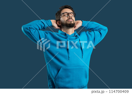 A young man wearing glasses and a casual blue hoodie stands with his hands behind his head, eyes closed in a gesture of relaxation and contentment, blue background 114700409