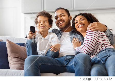 African American family of three, including two parents and child, is seated on a couch in a living room setting. They are holding a remote control, presumably watching television together. 114700412