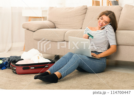 A young woman sitting on the floor of her living room, leaning against a sofa as she converses on her smartphone with a pleased smile, holding credit card and laptop, booking tickets or hotel A young woman sitting on the floor of her living room, leaning against a sofa as she converses on her smartphone with a pleased smile, holding credit card and laptop, booking tickets or hotel 114700447