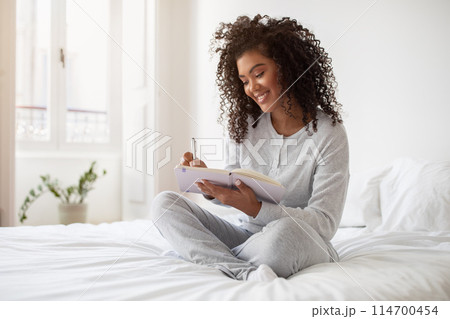 A Hispanic woman is seated on a bed, focused on writing in a notebook. She appears engrossed in her writing, pen in hand, surrounded by a cozy bedroom setting. A Hispanic woman is seated on a bed, focused on writing in a notebook. She appears engrossed in her writing, pen in hand, surrounded by a cozy bedroom setting. 114700454
