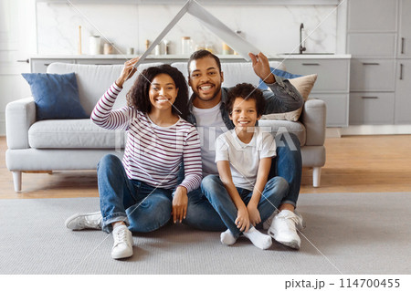A joyful African American family consisting of mother, father, and their young son is comfortably seated on the floor of their well-lit living room, holding paper cutout of house above their heads 114700455