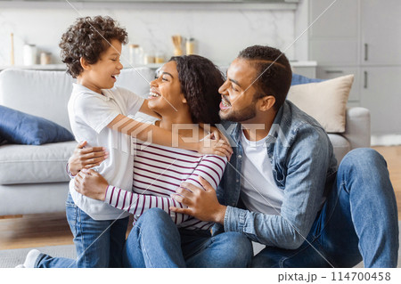African American man, woman, and child are seated on the floor in front of a couch. They appear to be engaged in conversation or possibly watching something together. African American man, woman, and child are seated on the floor in front of a couch. They appear to be engaged in conversation or possibly watching something together. 114700458