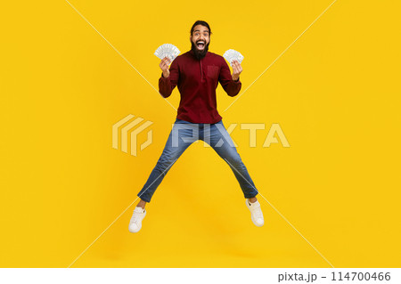 Indian man wearing a red sweater and blue jeans exuberates happiness while jumping in the air. He is holding a bunch of money in both hands, celebrating probably some sort of financial success 114700466