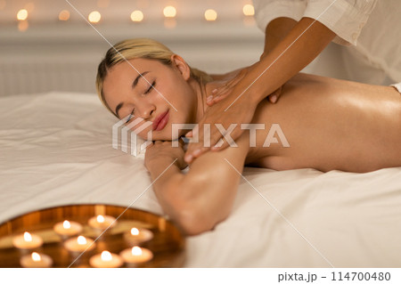 A serene spa scene where a young blonde woman enjoys a relaxing back massage, surrounded by a softly lit, calming environment. 114700480