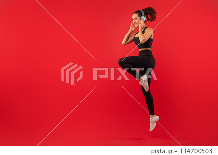 A joyful young woman with her hair tied in a ponytail is captured mid-jump against a bold red backdrop, showcasing her active lifestyle, using wireless headphones, copy space A joyful young woman with her hair tied in a ponytail is captured mid-jump against a bold red backdrop, showcasing her active lifestyle, using wireless headphones, copy space 114700503
