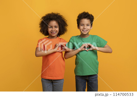 Happy African American kids boy and girl showing heart shape gestures over chest and smiling at camera, isolated on yellow studio background 114700533