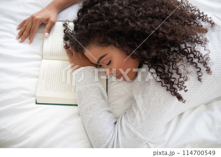 A young Hispanic woman with curly hair lies on a white bed, her head resting on an open book. She appears to be taking a break or perhaps has fallen asleep while reading 114700549