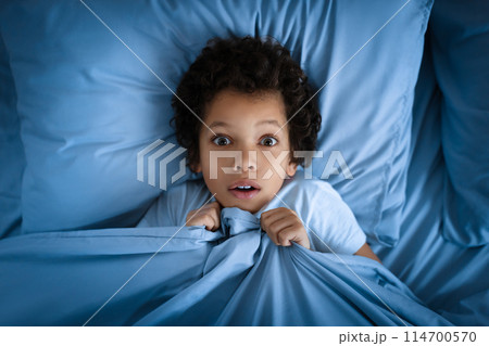 African American young boy lies in bed with a surprised look on his face, clutching the blue bed covers up to his chin. His wide eyes convey a sense of awe or alarm, top view 114700570