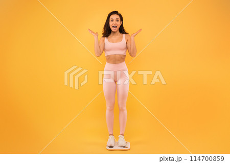 A young woman stands on a scale, expressing surprise and excitement with her hands raised. She is dressed in light-colored active wear and white sneakers, set against a vibrant yellow backdrop. A young woman stands on a scale, expressing surprise and excitement with her hands raised. She is dressed in light-colored active wear and white sneakers, set against a vibrant yellow backdrop. 114700859