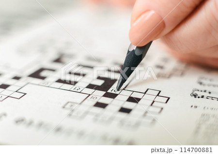 A person is seen actively engaged in a crossword puzzle, holding a pencil and carefully filling in the boxes with letters. The crossword grid is visible, showing a mix of empty and filled squares. 114700881