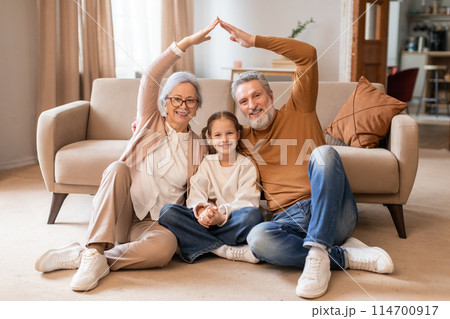 A smiling elderly couple and a young girl sit on the floor in a cozy living room, forming a house outline with their hands above them, symbolizing unity and family love. A smiling elderly couple and a young girl sit on the floor in a cozy living room, forming a house outline with their hands above them, symbolizing unity and family love. 114700917