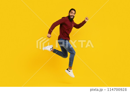 Indian man wearing a red sweater is captured mid-jump in the air. His arms are raised, and his legs are bent as he springs upwards. The background is yellow, emphasizing the mans movement. Indian man wearing a red sweater is captured mid-jump in the air. His arms are raised, and his legs are bent as he springs upwards. The background is yellow, emphasizing the mans movement. 114700928