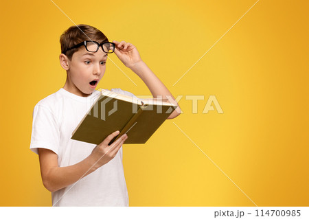A young boy wearing glasses is engrossed in reading a book. 114700985