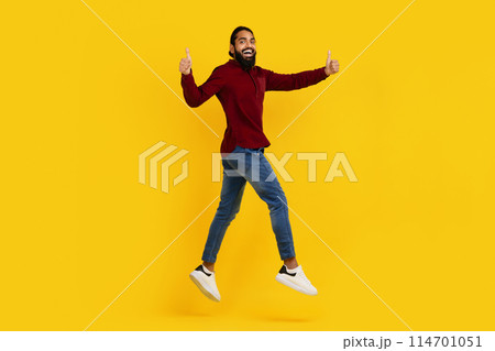 Indian man wearing casual clothing is captured mid-jump with his arms lifted towards the sky, showing thumbs up isolated on yellow studio background 114701051