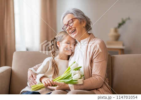 A cheerful elderly woman with glasses is warmly hugging her young granddaughter, who holds a bouquet of fresh white tulips, home interior 114701054